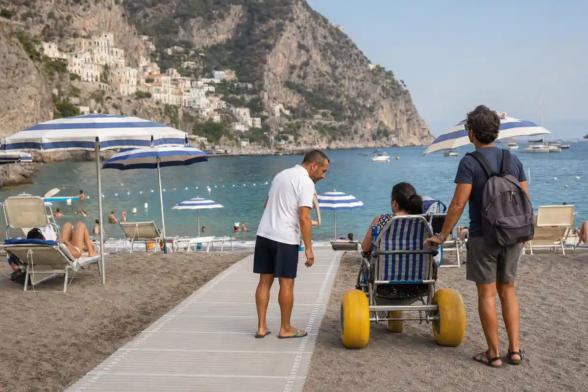 Passerella accessibile in spiaggia con sedia da mare a ruote e assistente, ombrelloni e mare sullo sfondo