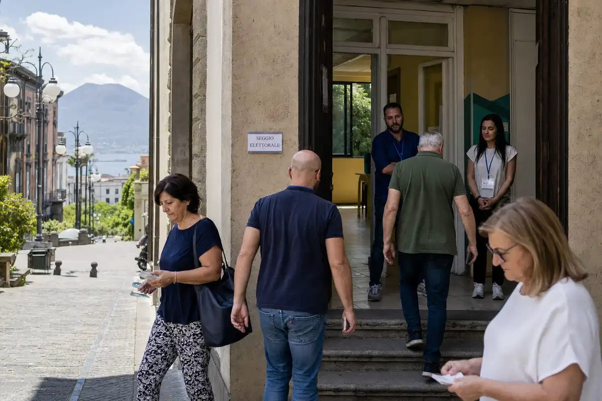 Elettori entrano in un seggio elettorale con personale all’ingresso, con vista su una strada e un vulcano sullo sfondo