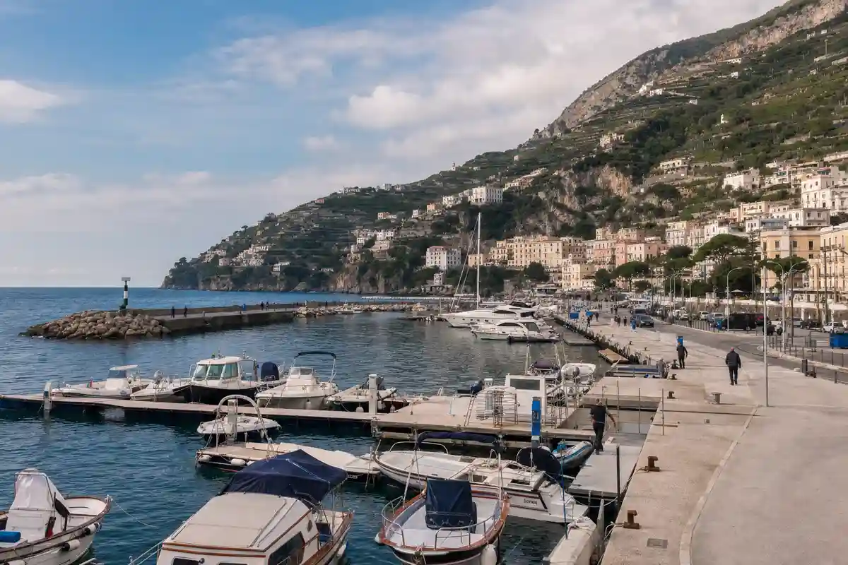 Porticciolo con barche e piccoli yacht ormeggiati, molo e frangiflutti, con borgo costiero e colline terrazzate sullo sfondo